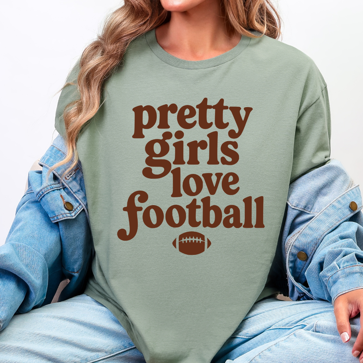 Woman wearing a sage Pretty Girls Love Football graphic tee with brown retro lettering and a football design.