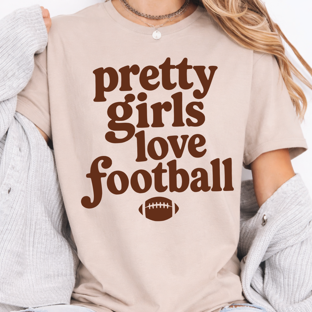 Woman wearing a sand Pretty Girls Love Football graphic tee with brown retro lettering and a football design.