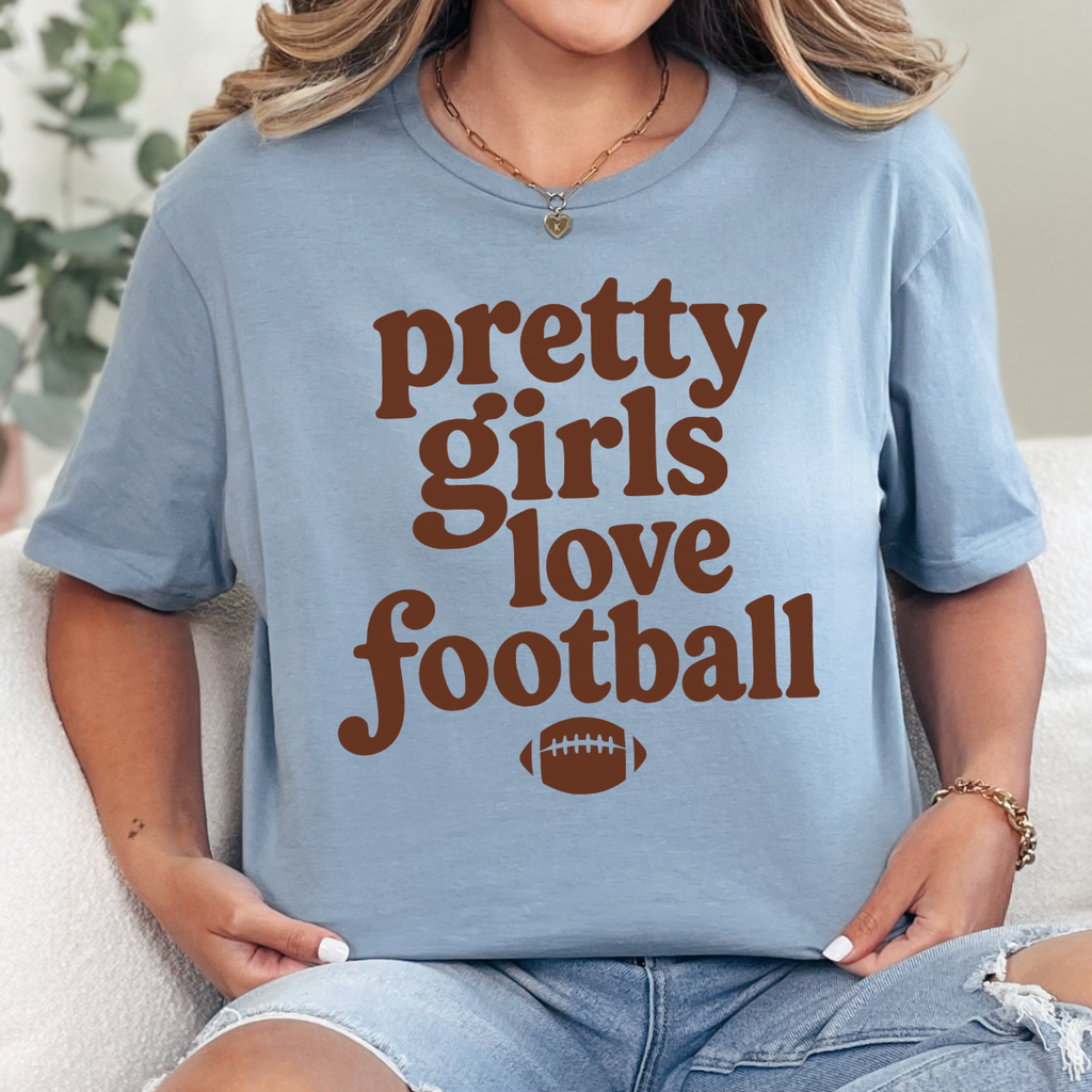 Woman wearing a stone blue Pretty Girls Love Football graphic tee with brown retro lettering and a football design.