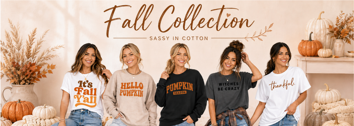 Five women seated together wearing fall-themed graphic t-shirts with autumn quotes like 'It's Fall You', 'Hello Pumpkin', and 'Pumpkin Spice', posed in a decorated indoor setting with pumpkins and the text 'Fall Collection' at the top.