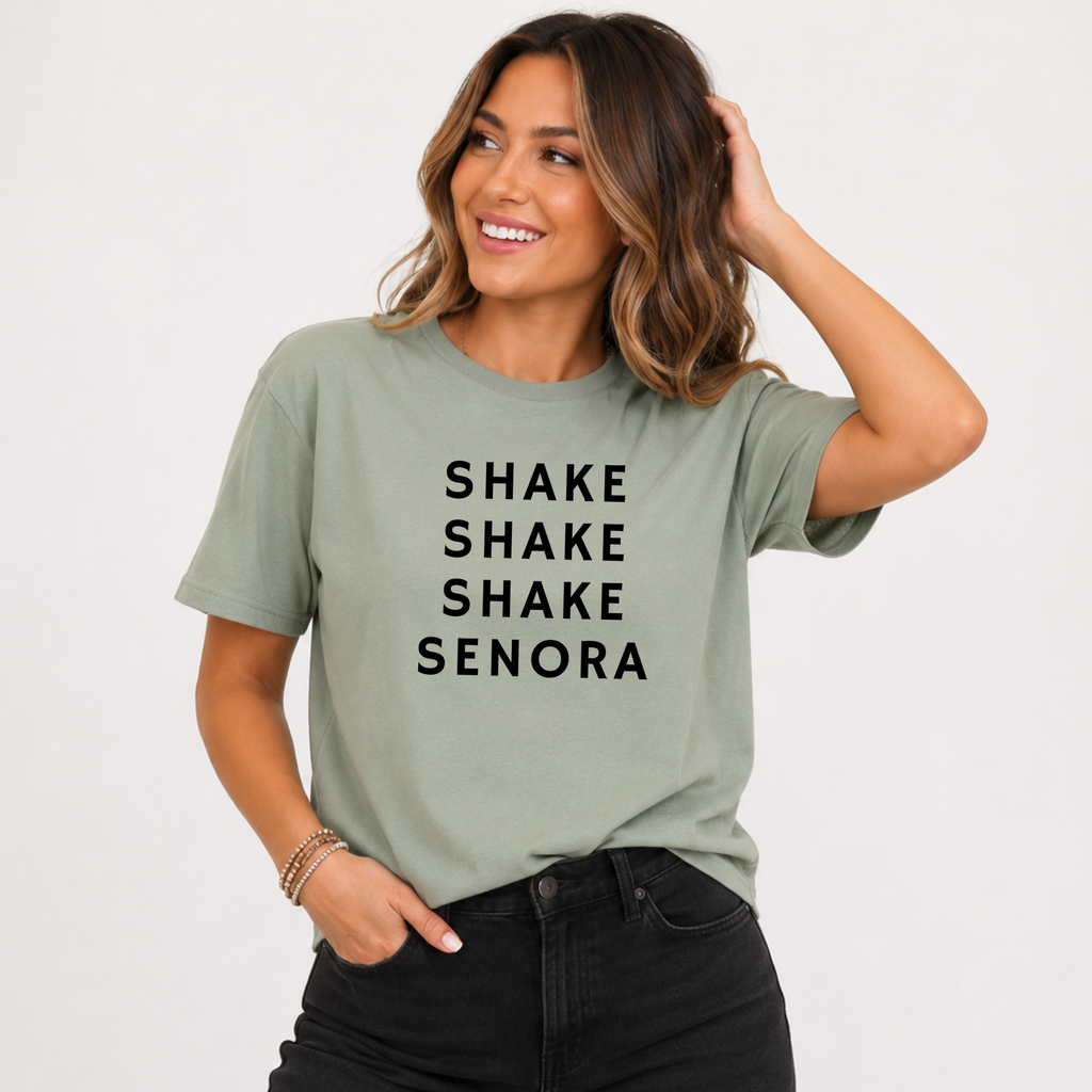Woman wearing a sage green Shake Shake Shake Señora Halloween graphic tee with black lettering.