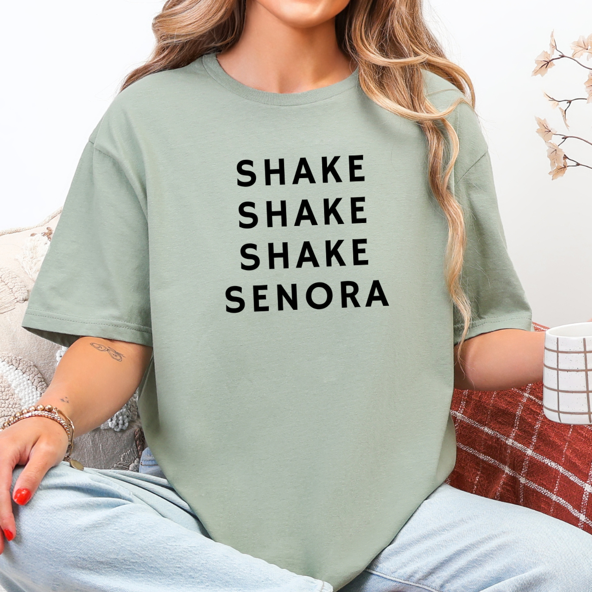 Woman wearing a sage green Shake Shake Shake Señora Halloween graphic tee with black lettering.