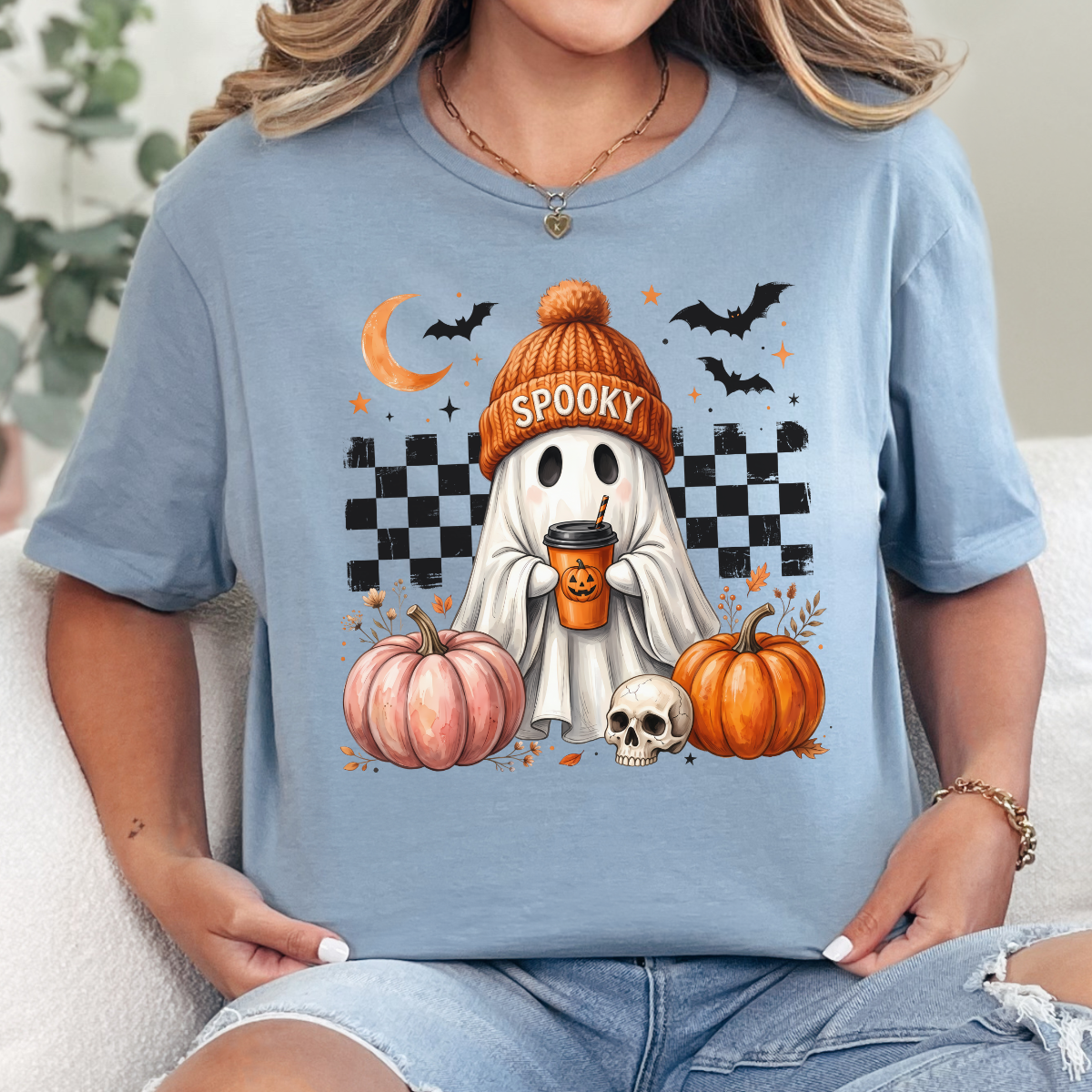 Woman wearing a blue spooky ghost pumpkin coffee graphic tee with pumpkins, bats, and a ghost in an orange beanie