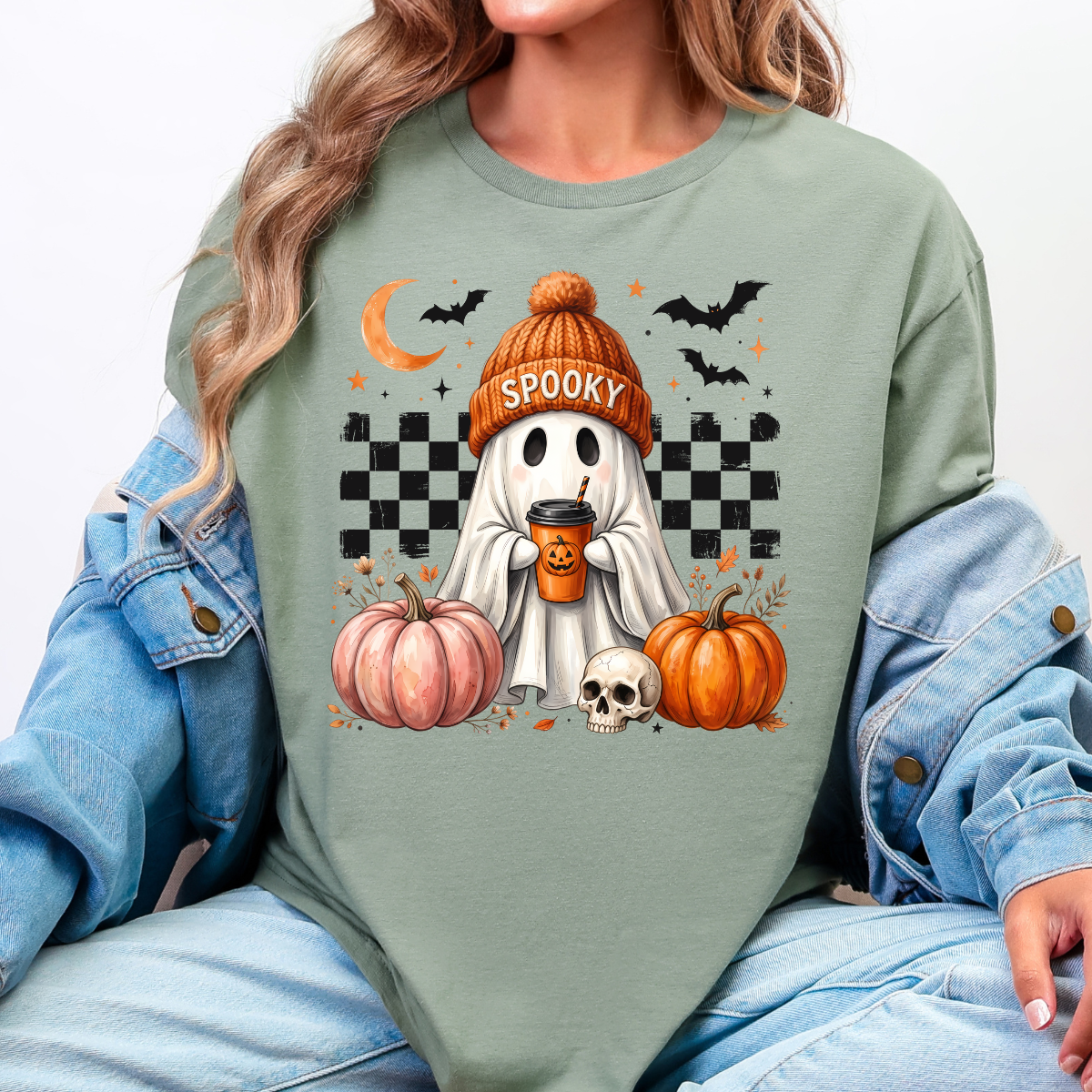 Woman wearing a sage green spooky ghost pumpkin coffee graphic tee with pumpkins, bats, and a ghost in an orange beanie