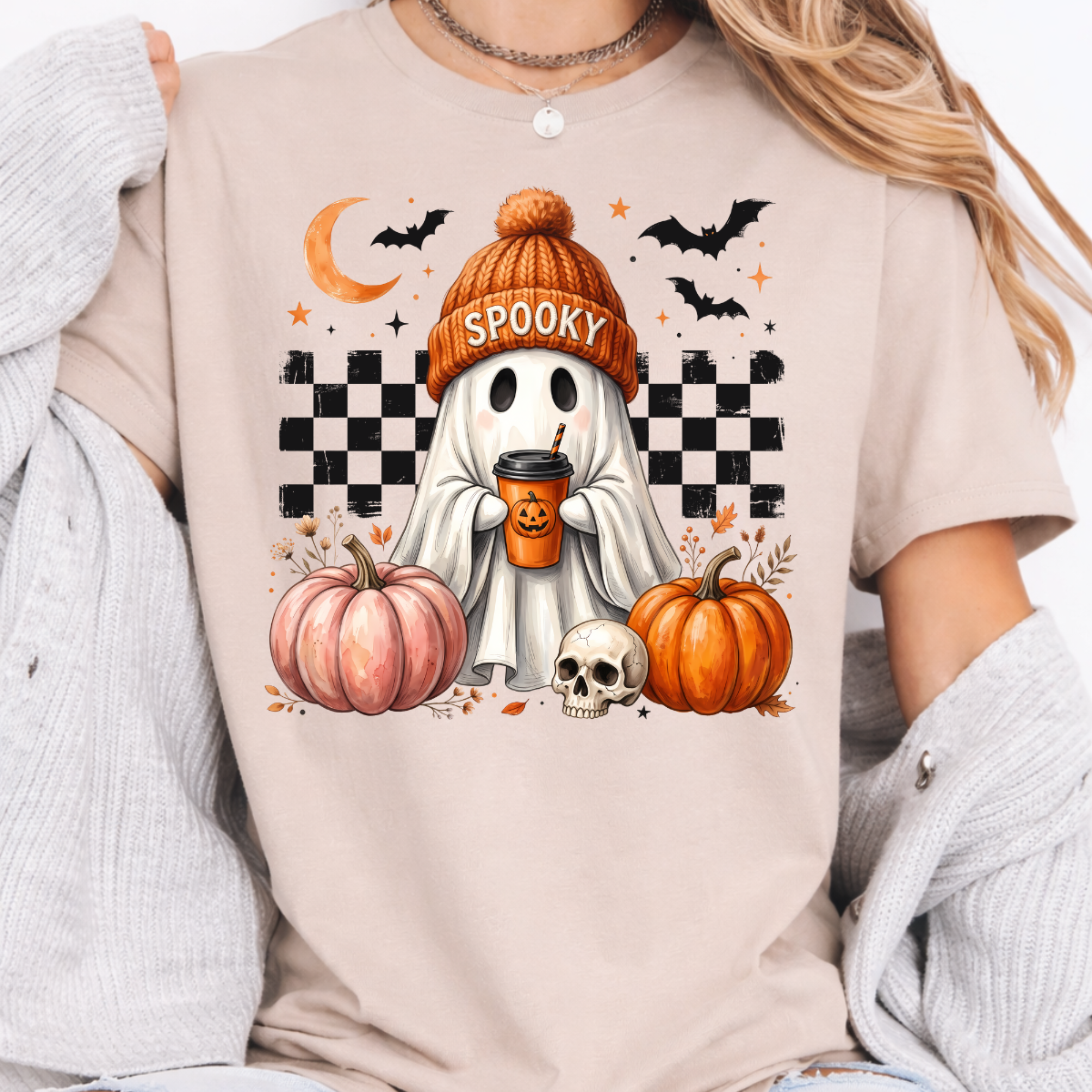 Woman wearing a cream sand spooky ghost pumpkin coffee graphic tee with pumpkins, bats, and a ghost.