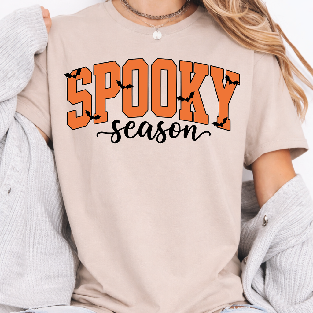 Woman wearing a sand Spooky Season Halloween graphic tee with orange lettering and black bats.