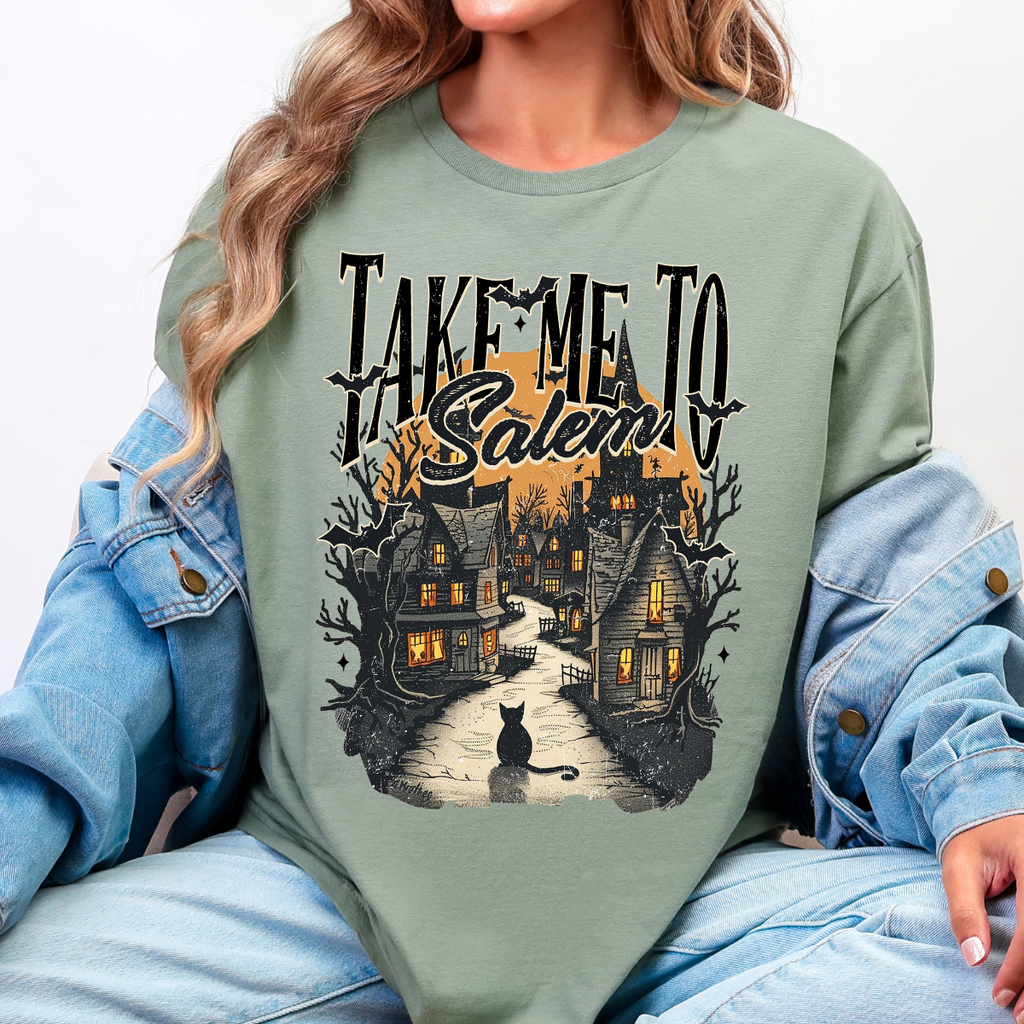 Woman wearing a Take Me to Salem Halloween graphic tee with a haunted village, black cat, bats, and orange moon.