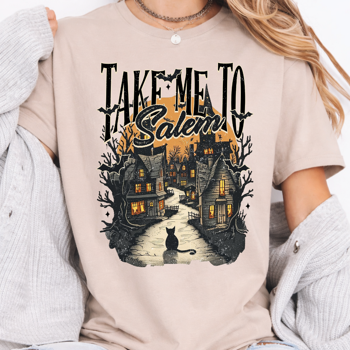 Woman wearing a Take Me to Salem Halloween graphic tee with a haunted village, black cat, bats, and orange moon.