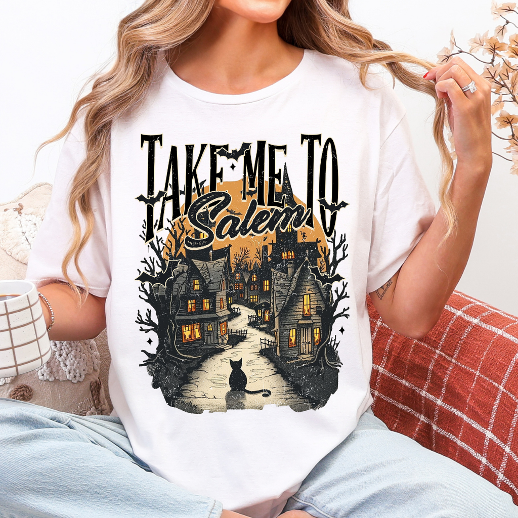 Woman wearing a Take Me to Salem Halloween graphic tee with a haunted village, black cat, bats, and orange moon.