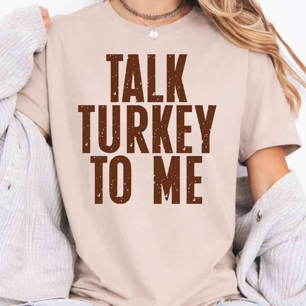 Woman wearing a sand Talk Turkey to Me Thanksgiving graphic tee with distressed brown lettering