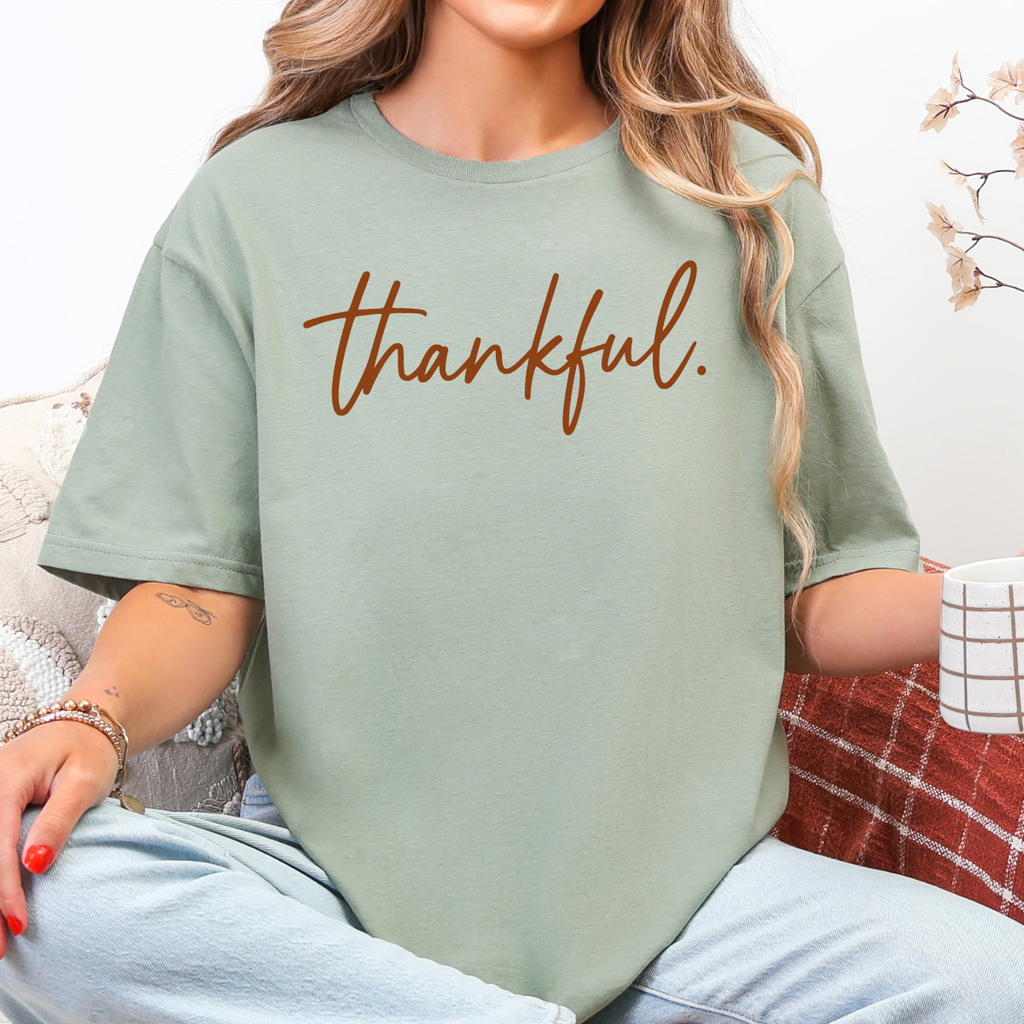 Woman wearing a sage green Thankful Thanksgiving graphic tee with warm brown script lettering.