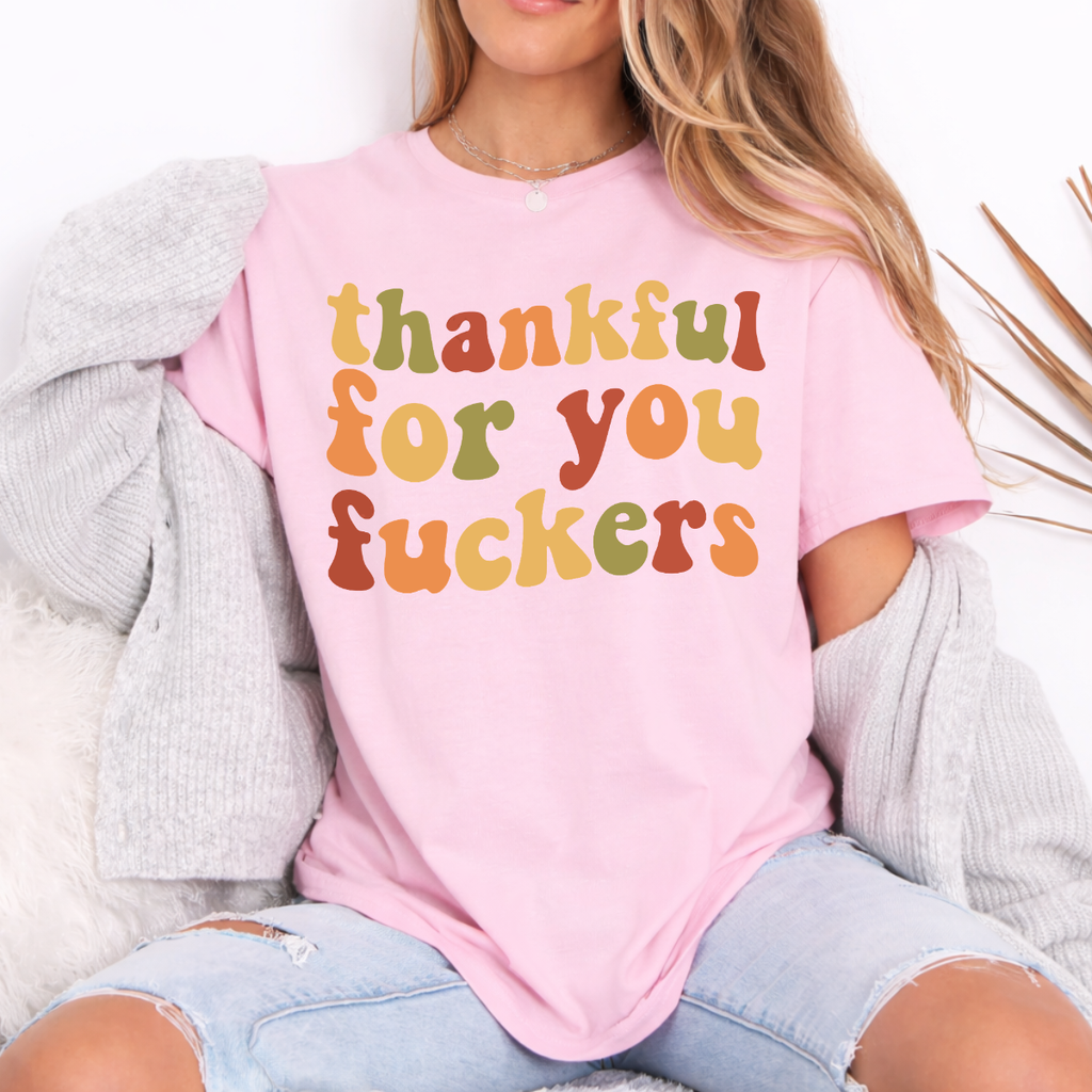 Woman wearing a light pink Thankful for You Fuckers Thanksgiving graphic tee with colorful retro lettering.