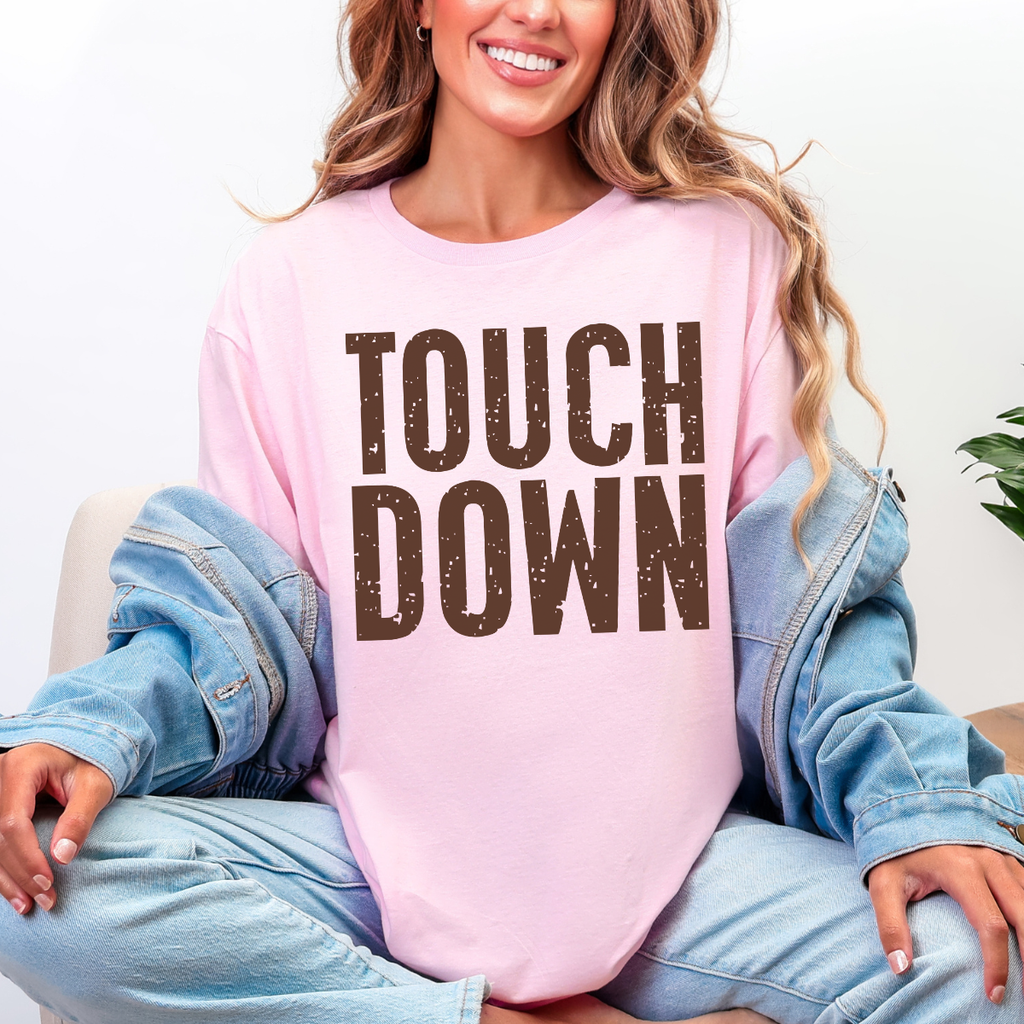 Woman wearing a cream Touchdown football graphic tee with bold brown distressed lettering.