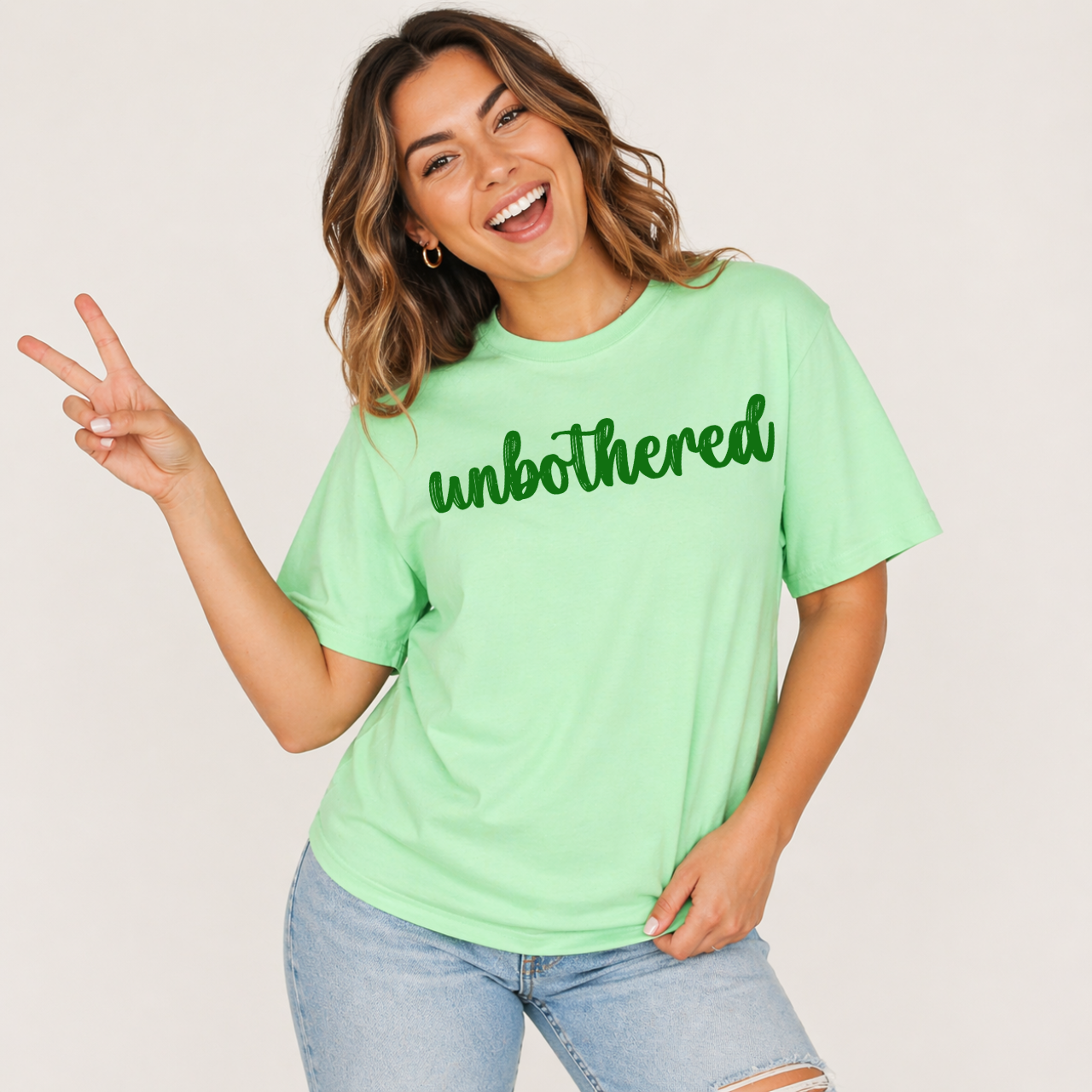 Woman wearing a light green t-shirt with 'unbothered' text, making a peace sign.