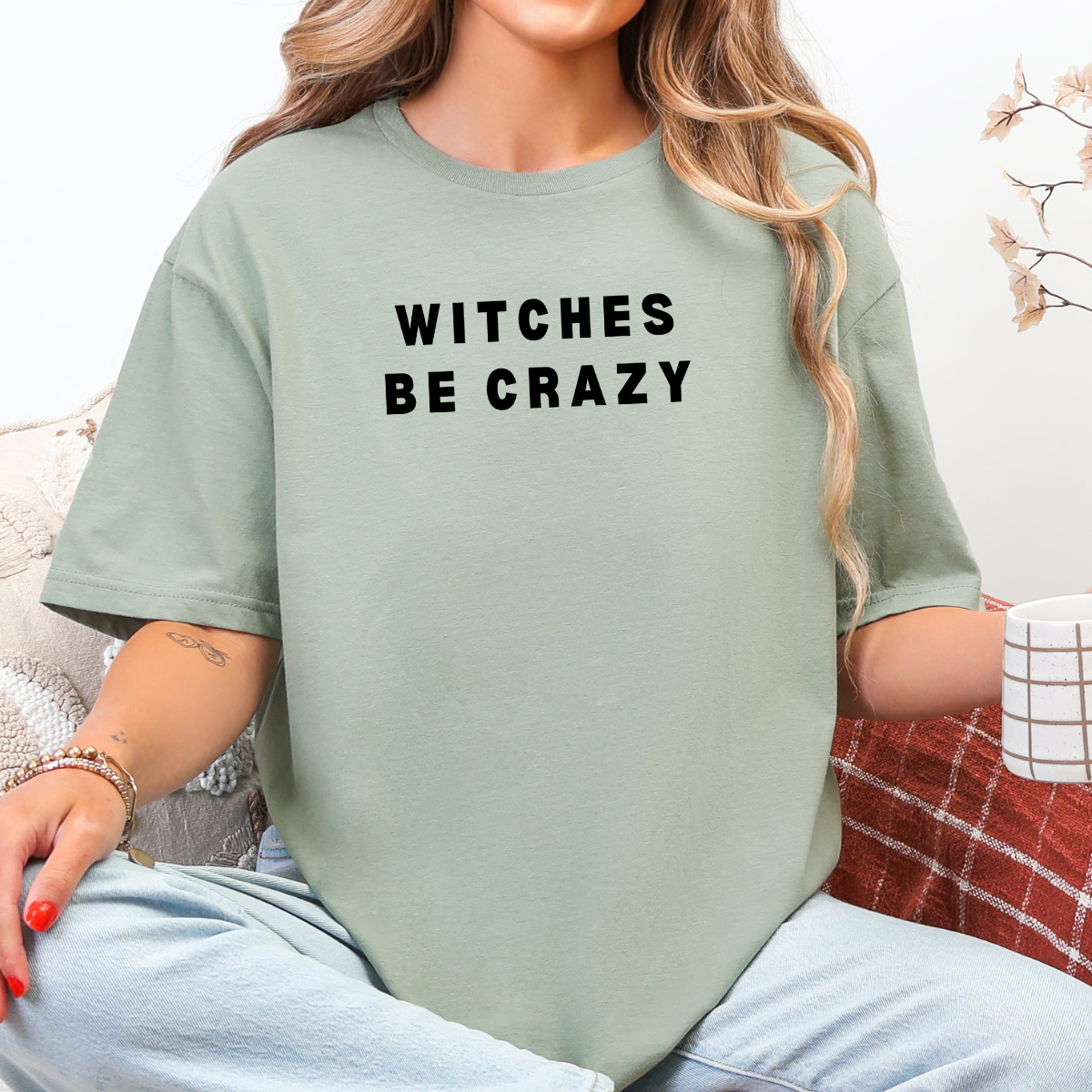 Woman wearing a sage Witches Be Crazy Halloween graphic tee with black lettering.