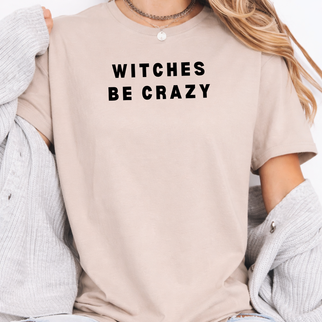 Woman wearing a sand Witches Be Crazy Halloween graphic tee with black lettering.